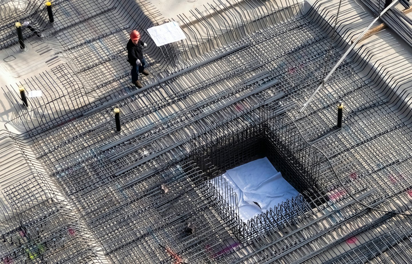 Construction worker inspects blueprints on rebar grid foundation.